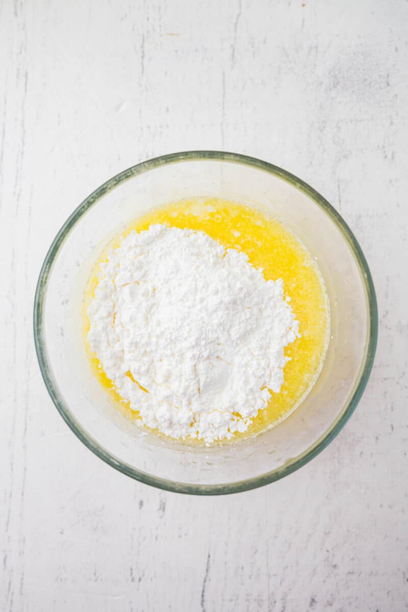 A glass bowl containing melted butter and a mound of white powdered sugar on a white surface.