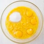 A glass bowl containing mashed bananas, four raw eggs, and a mound of sugar on a white surface.