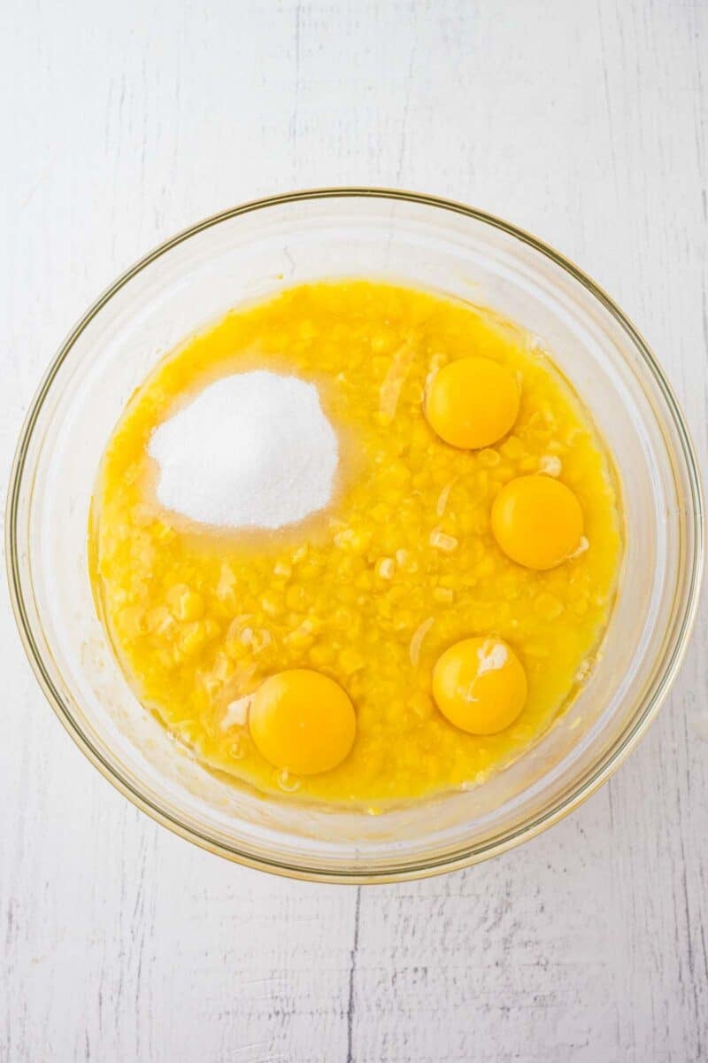 A glass bowl containing mashed bananas, four raw eggs, and a mound of sugar on a white surface.
