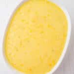 A white oval dish filled with a smooth, yellow batter containing small lumps, placed on a white wooden surface.