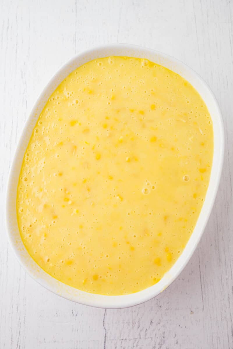 A white oval dish filled with a smooth, yellow batter containing small lumps, placed on a white wooden surface.