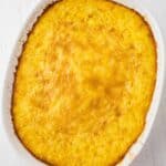 A baked casserole dish filled with a golden-brown cornbread pudding, shown from above on a white surface.