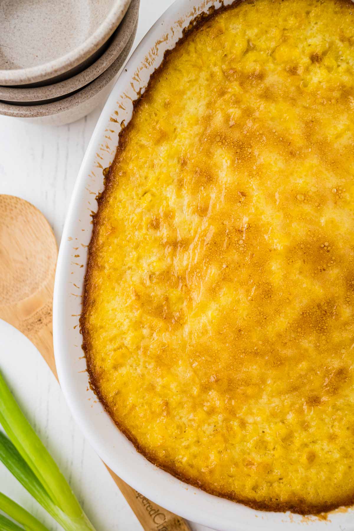 A baked corn pudding in a white casserole dish next to stacked plates, wooden utensils, and green onions on a white surface.