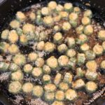Sliced okra pieces coated in cornmeal fry in oil in a black cast iron skillet on a stovetop.
