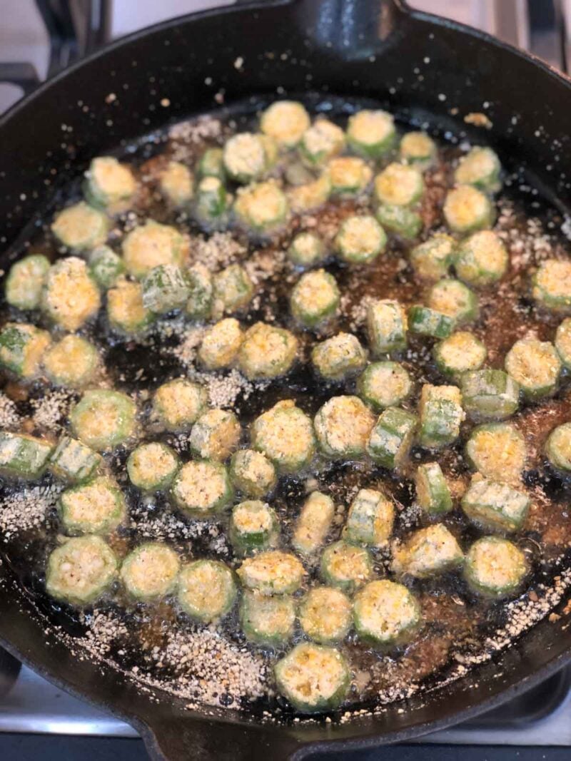 Sliced okra pieces coated in cornmeal fry in oil in a black cast iron skillet on a stovetop.