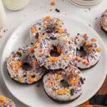 A plate of chocolate donuts with white icing and colorful Halloween-themed sprinkles, surrounded by bottles of milk and an orange and black plaid napkin.