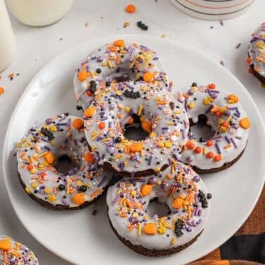 A plate of chocolate donuts with white icing and colorful Halloween-themed sprinkles, surrounded by bottles of milk and an orange and black plaid napkin.