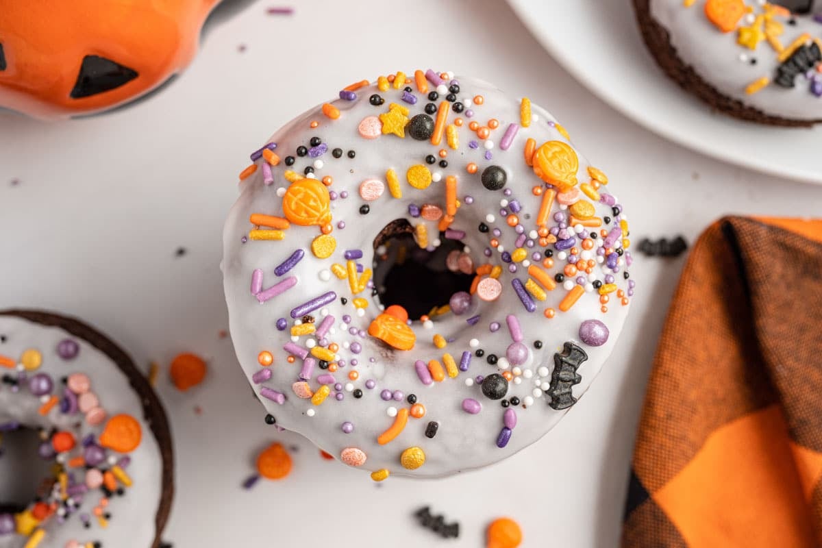 A chocolate donut with white icing topped with colorful Halloween-themed sprinkles, surrounded by festive decorations.