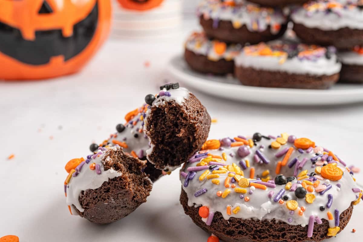 Chocolate donuts with white icing and colorful Halloween-themed sprinkles, one with a bite taken out, are displayed on a white surface with a jack-o'-lantern decoration in the background.