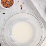 A glass bowl with white icing, a small bowl of Halloween-themed sprinkles, a chocolate donut, and a white spatula on a gray surface.