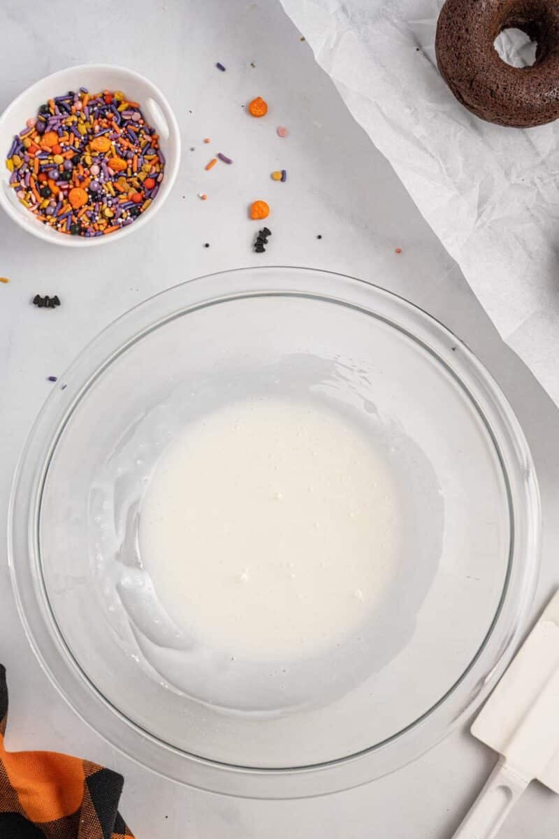 A glass bowl with white icing, a small bowl of Halloween-themed sprinkles, a chocolate donut, and a white spatula on a gray surface.