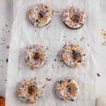 Six chocolate donuts with purple, orange, and black sprinkles are arranged on parchment paper, with a bowl of sprinkles and a jack-o'-lantern decoration nearby.