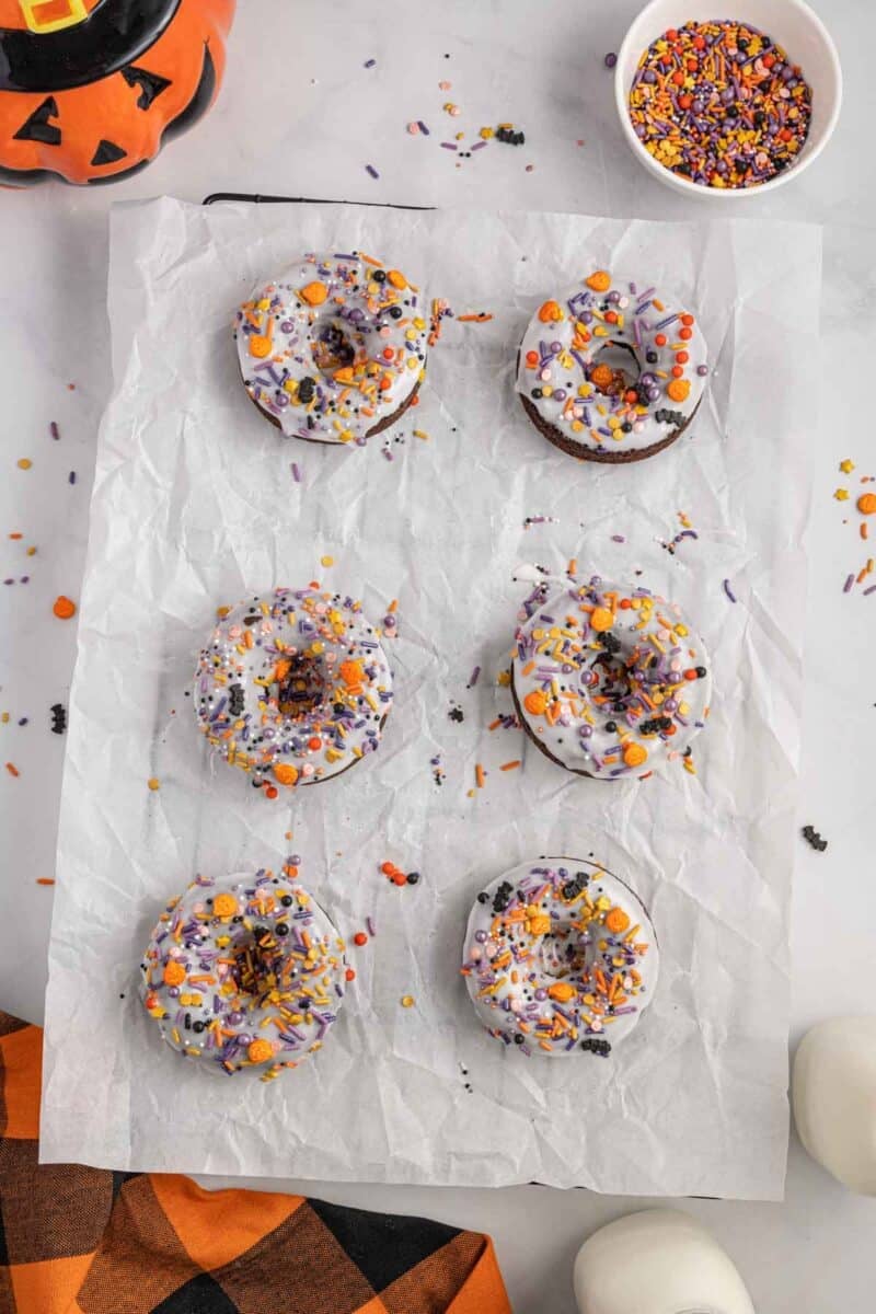 Six chocolate donuts with purple, orange, and black sprinkles are arranged on parchment paper, with a bowl of sprinkles and a jack-o'-lantern decoration nearby.
