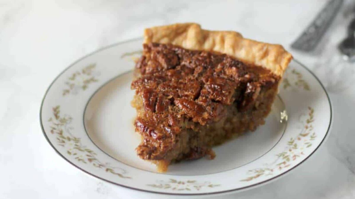 A slice of pecan pie, one of the ultimate comfort desserts, sits on a decorative white plate with a gold rim, set on a light surface.