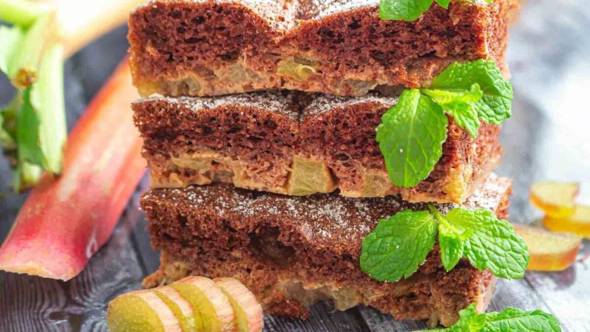 Three slices of chocolate and rhubarb cake are stacked on a wooden surface, garnished with fresh mint and powdered sugar, with rhubarb pieces nearby—a delicious addition to your collection of indulgent desserts.