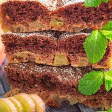Three slices of chocolate and rhubarb cake are stacked on a wooden surface, garnished with fresh mint and powdered sugar, with rhubarb pieces nearbyโa delicious addition to your collection of indulgent desserts.