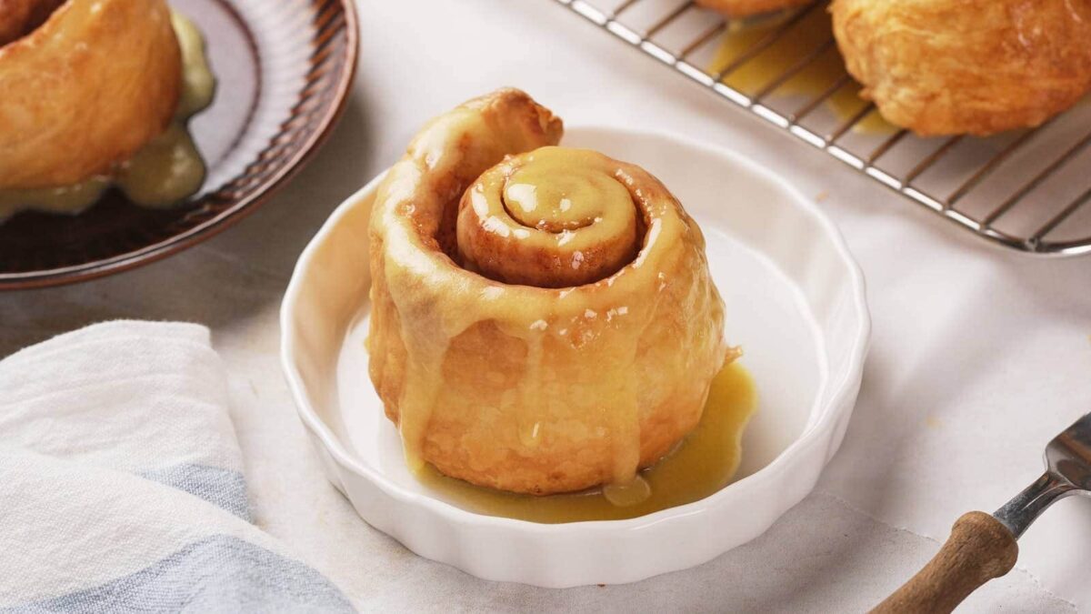 A cinnamon roll topped with icing sits on a white dish; more cinnamon rolls and a cooling rack are visible in the background, showcasing this delicious cinnamon rolls recipe.