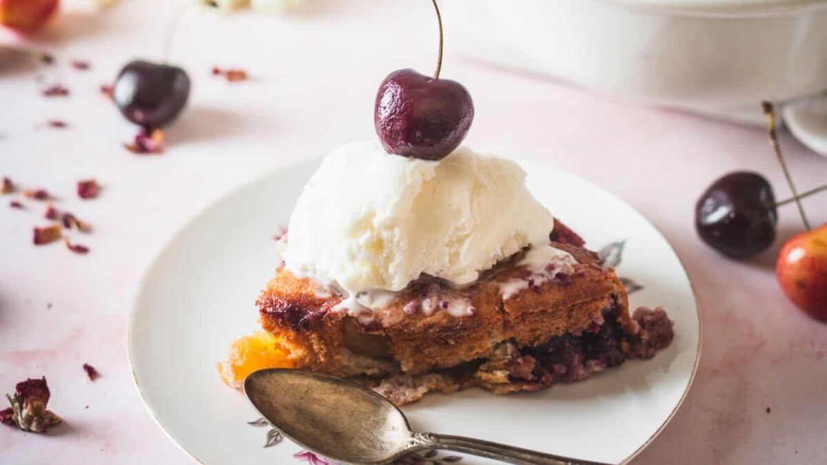 A slice of cherry cobbler topped with vanilla ice cream and a cherry, served on a white plate with a spoon—one of the best easy cherry desserts for sweet cravings.