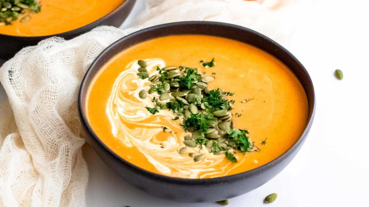 A bowl of creamy orange soup with a swirl of cream, topped with chopped parsley and pumpkin seeds, delivers big flavor—perfect for fans of easy curry and creative curry recipes. Served on a white surface.