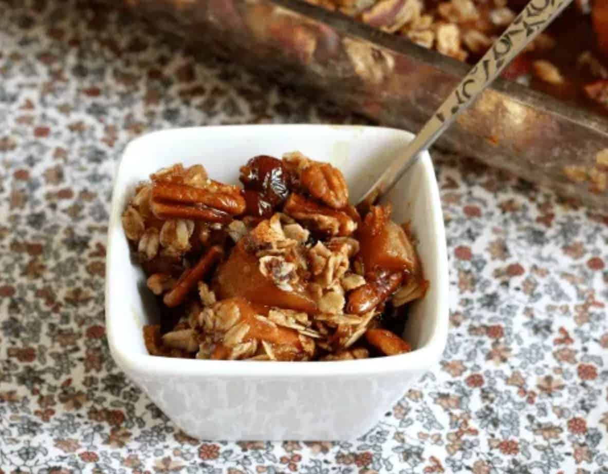 A small white bowl filled with granola, nuts, raisins, and apple chunks makes for a wholesome gluten-free treat; a larger dish of the same food is in the background, with a spoon resting inside.