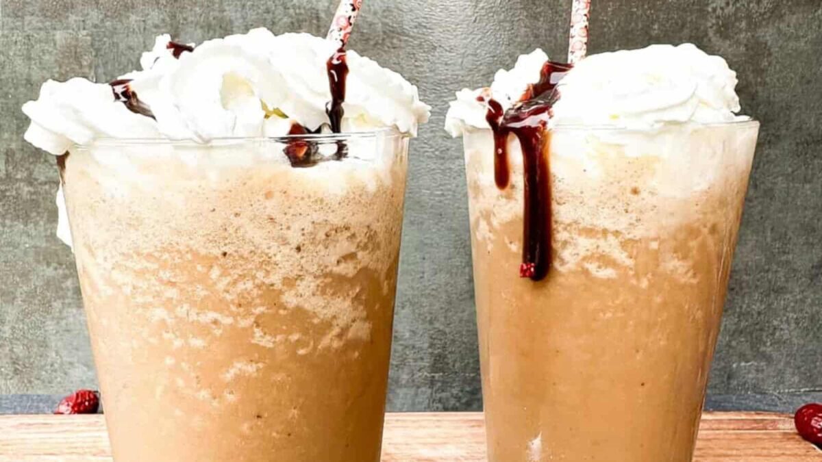 Two glasses of indulgent gluten-free iced coffee topped with whipped cream and chocolate syrup, each with a straw, sit on a wooden surface against a gray background.