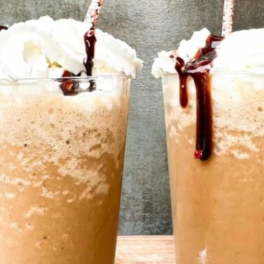 Two glasses of indulgent gluten-free iced coffee topped with whipped cream and chocolate syrup, each with a straw, sit on a wooden surface against a gray background.