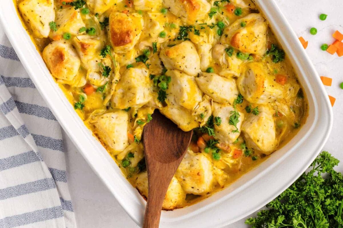 A casserole dish filled with creamy chicken and the best biscuits, mixed with peas and carrots, with a wooden spoon scooping from it.