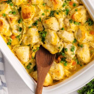 A casserole dish filled with creamy chicken and the best biscuits, mixed with peas and carrots, with a wooden spoon scooping from it.