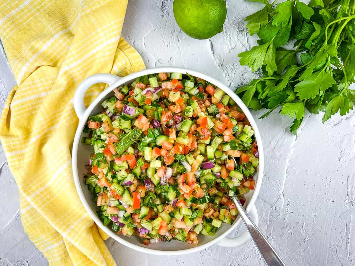 A white bowl filled with chopped salad—cucumber, tomato, red onion, and herbs—offers a sunny escape into Mediterranean cuisine. A spoon is in the bowl, with fresh parsley, a lime, and a yellow towel nearby.