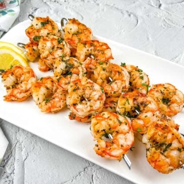 Three metal skewers with grilled shrimp garnished with herbs are arranged on a white plate with lemon wedges on the side, capturing the flavors of Mediterranean cuisine for a sunny escape at your table.
