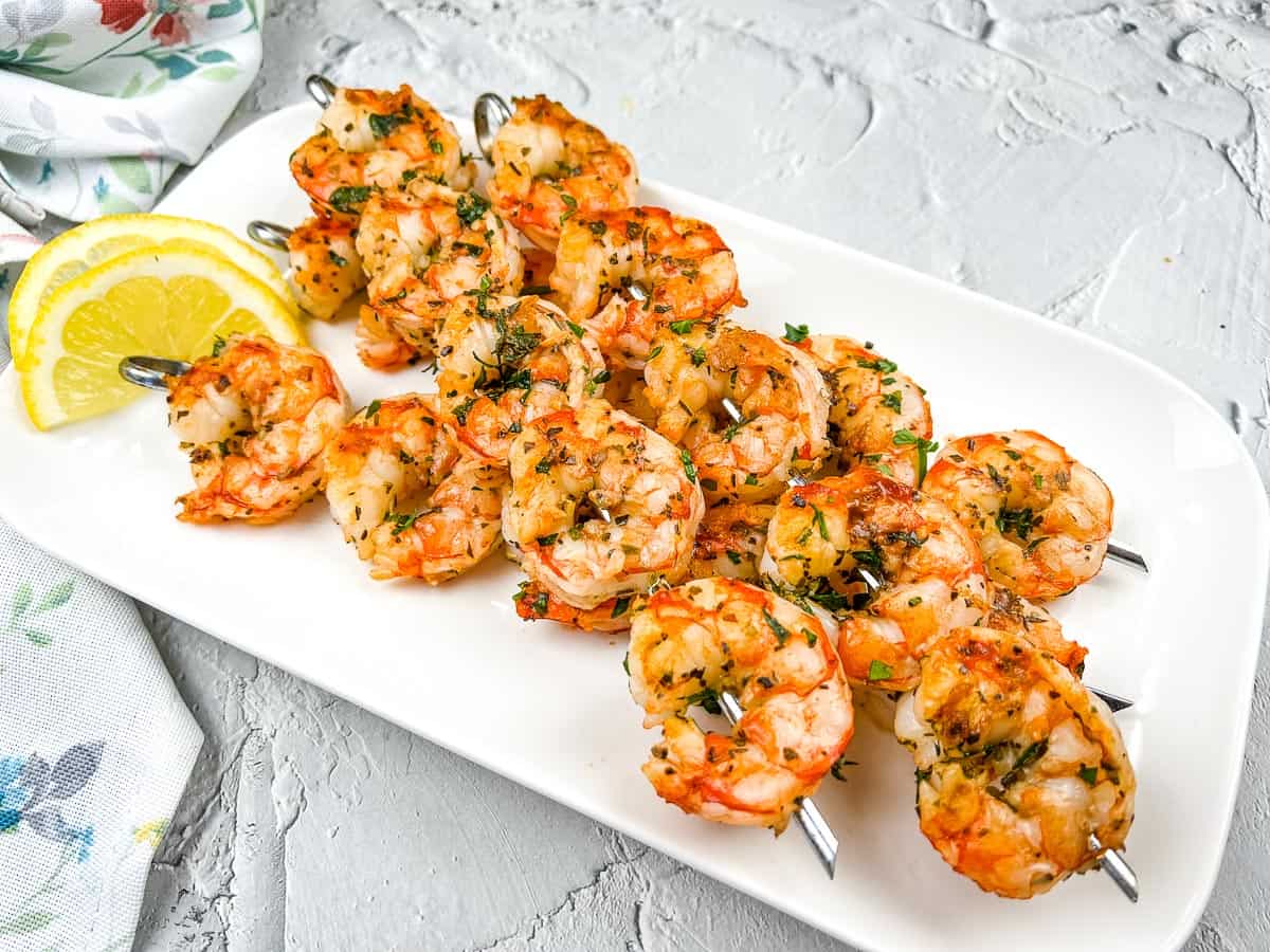 Three metal skewers with grilled shrimp garnished with herbs are arranged on a white plate with lemon wedges on the side, capturing the flavors of Mediterranean cuisine for a sunny escape at your table.