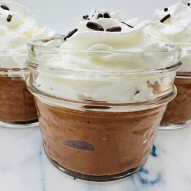 Three glass jars filled with chocolate mousse, topped with whipped cream and chocolate sprinkles, are displayed on a white marble surface—perfect for lovers of chocolate desserts or celebration desserts.