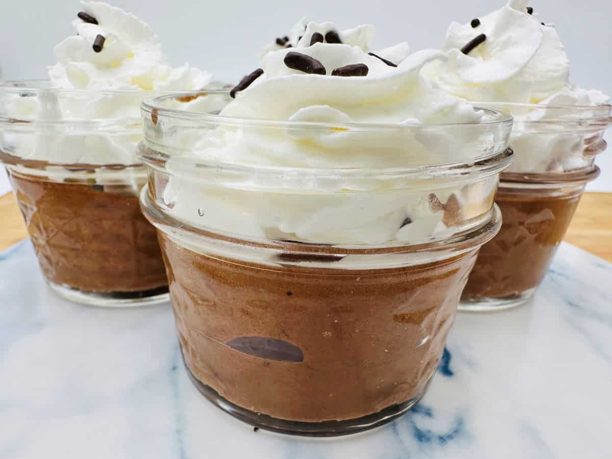 Three glass jars filled with chocolate mousse, topped with whipped cream and chocolate sprinkles, are displayed on a white marble surface—perfect for lovers of chocolate desserts or celebration desserts.