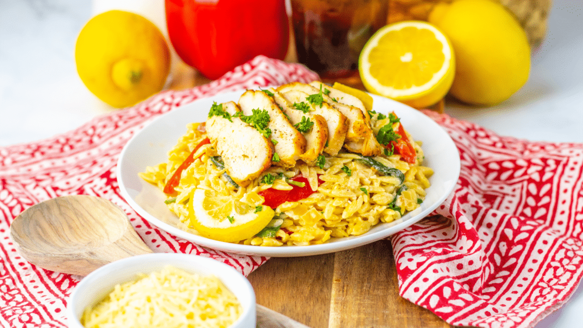 A plate of creamy orzo pasta with sliced grilled chicken, red peppers, spinach, and lemon garnish sits on a wooden board—perfect for budget dinners, with grated cheese and fresh vegetables nearby.