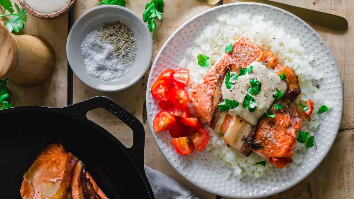 A plate of cooked salmon fillets topped with melted cheese and parsley, served over rice with sliced cherry tomatoes on the side—the perfect addition to your party recipes. Salt, pepper, and herbs are nearby.