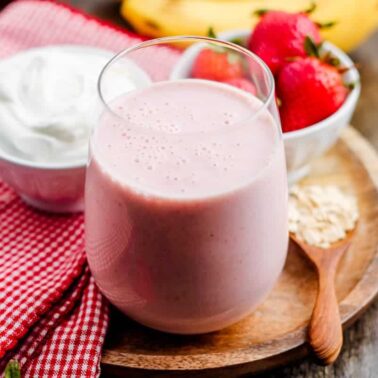 A glass of delicious pink smoothie on a wooden plate with a bowl of yogurt, strawberries, a wooden spoon with oats, and a red-checkered cloth nearby—perfect inspiration for healthy smoothie recipes.