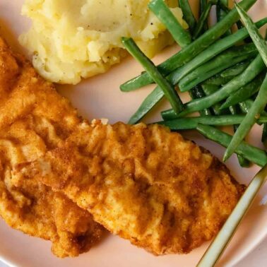 A plate with two breaded chicken tenders, mashed potatoes, green beans, and a gold fork—perfect for enjoying homemade takeout or trying your hand at copycat dishes.