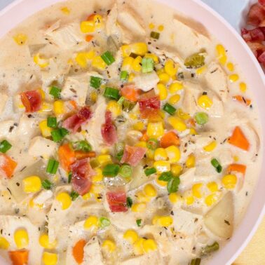 A bowl of creamy chicken and corn chowder, a true home favorite, topped with chopped green onions and bacon, with extra bacon and green onions in small bowls on the side.