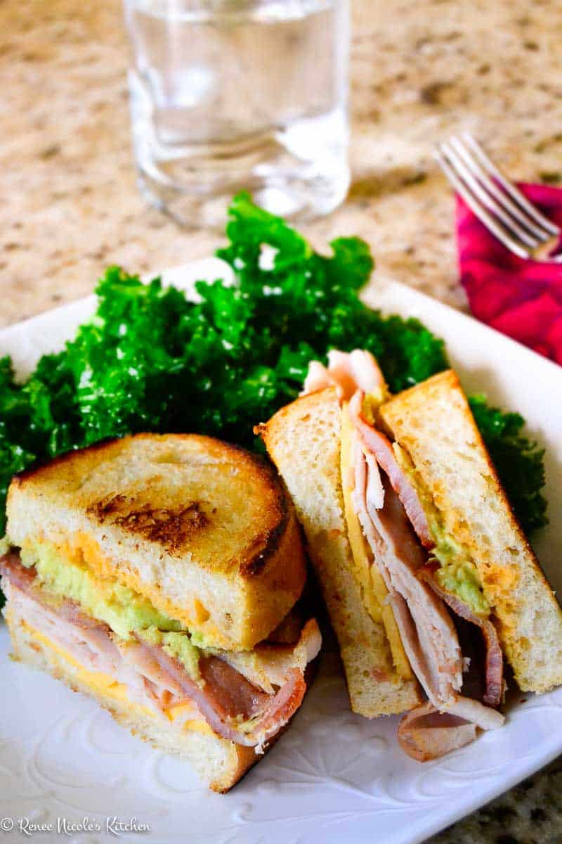 A delicious lunch option, this sandwich with turkey, bacon, and avocado is cut in half on a white plate beside leafy green kale—a tasty addition to your list of sandwich ideas. A glass of water and a fork sit in the background.