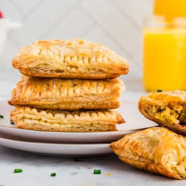 Four golden puff pastry breakfast pockets are stacked on a plate, one cut open to reveal a scrambled egg filling—an enjoyable, kid-friendly recipe perfect for mealtime. Two glasses of orange juice and berries sit in the background.