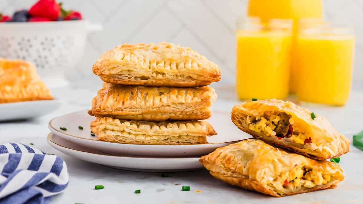 Four golden puff pastry breakfast pockets are stacked on a plate, one cut open to reveal a scrambled egg filling—an enjoyable, kid-friendly recipe perfect for mealtime. Two glasses of orange juice and berries sit in the background.
