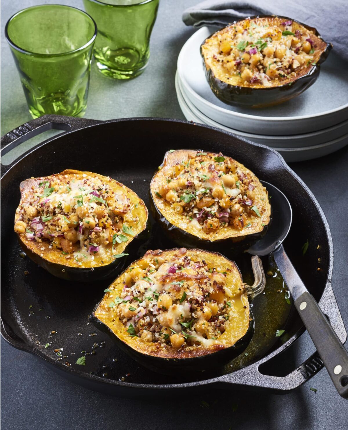 Three halves of stuffed acorn squash in a cast iron skillet, topped with herbs and cheese—this cozy dish is perfect for vegetarian meals, with two green glasses and stacked plates adding charm in the background.