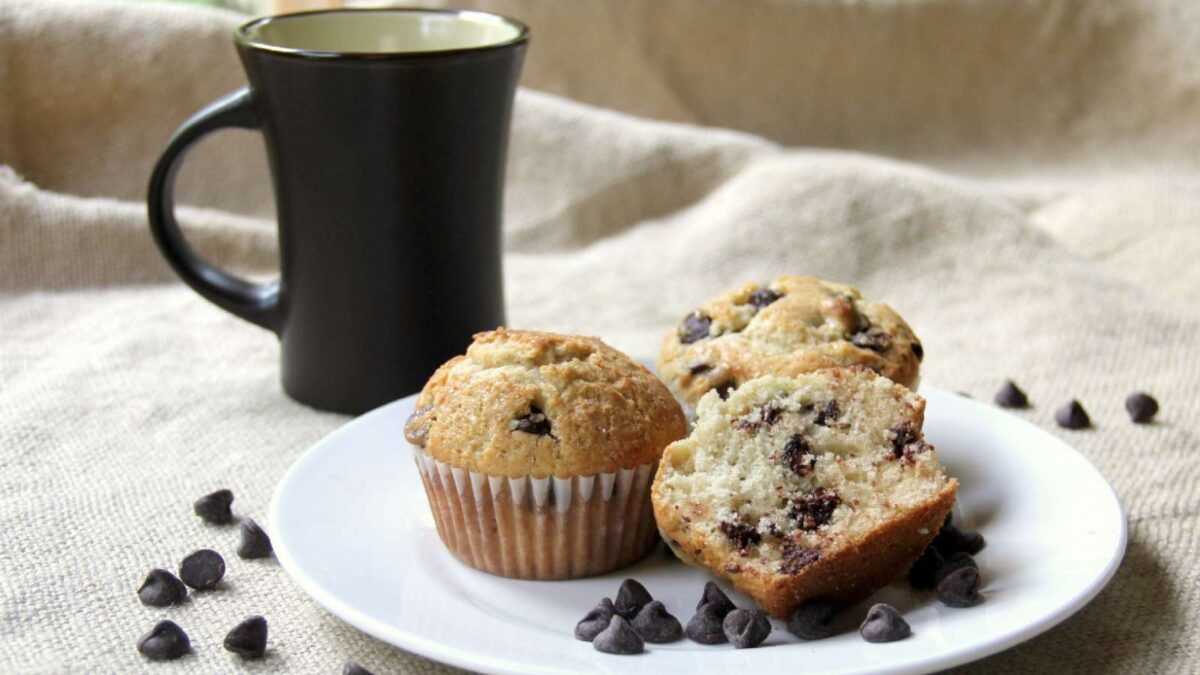A white plate with two chocolate chip muffins—one whole, one cut in half—surrounded by chocolate chips, next to a dark coffee mug. Discover unique muffins with unexpected ingredients in your muffin recipes.