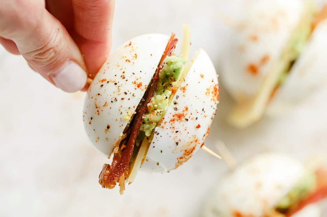 A hand holds a halved hard-boiled egg filled with avocado, cheese, and bacon, seasoned with pepper and spices, secured with a toothpick—a tasty twist for bacon recipes lovers.