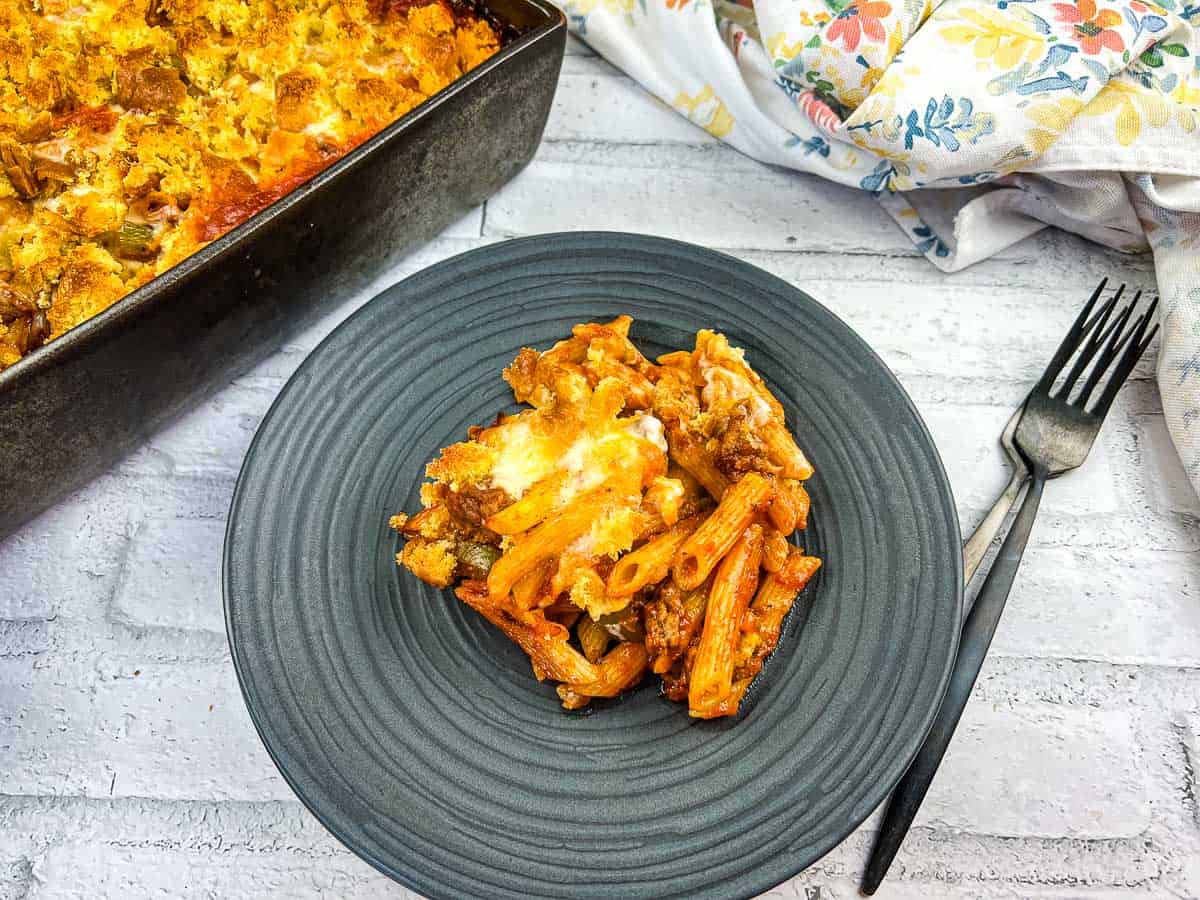 A serving of baked ziti with melted cheese on a gray plate, next to a black fork and a pan of the remaining casserole—perfect for stress-free meals that easily feed crowds.