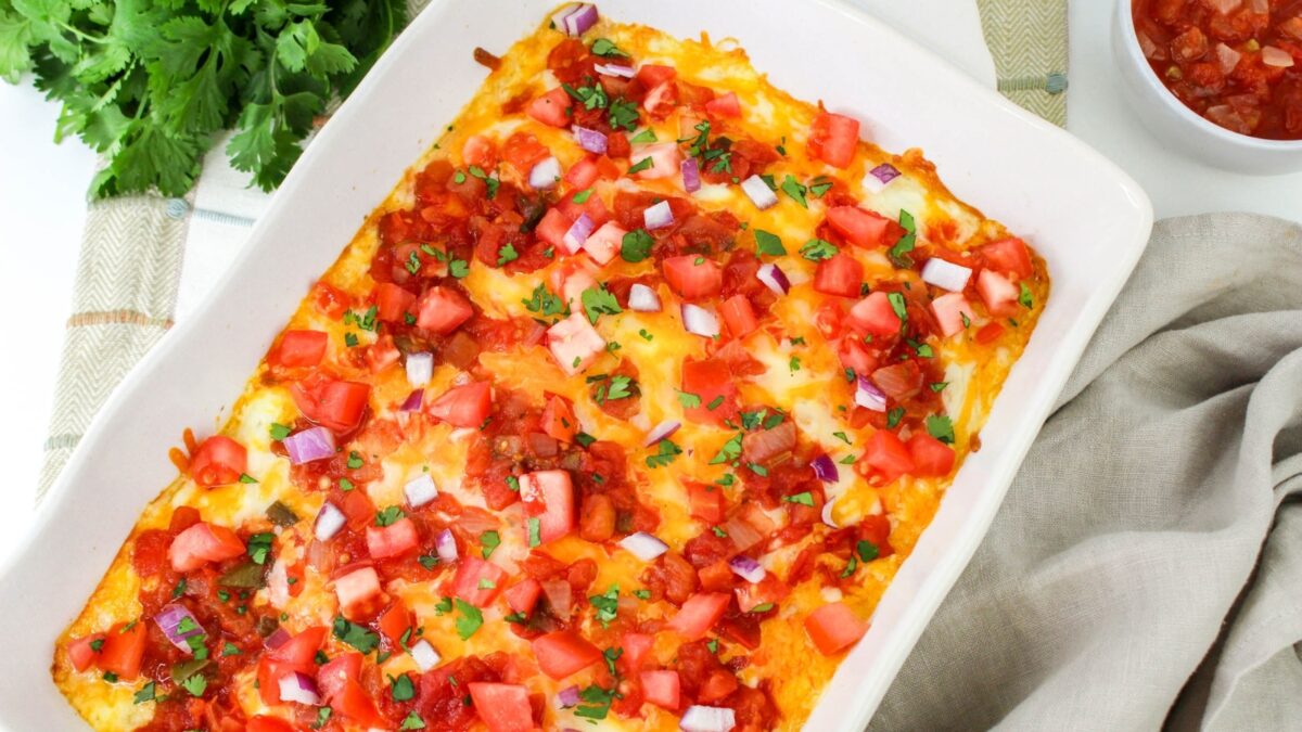 A white baking dish filled with a baked casserole topped with melted cheese, diced tomatoes, red onions, and chopped herbs sits on a table next to a bowl of salsa and fresh cilantro—perfect for easy casseroles to feed a crowd.
