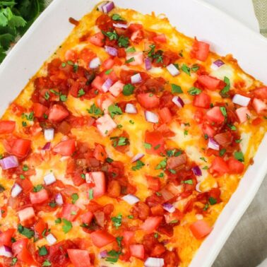 A white baking dish filled with a baked casserole topped with melted cheese, diced tomatoes, red onions, and chopped herbs sits on a table next to a bowl of salsa and fresh cilantro—perfect for easy casseroles to feed a crowd.