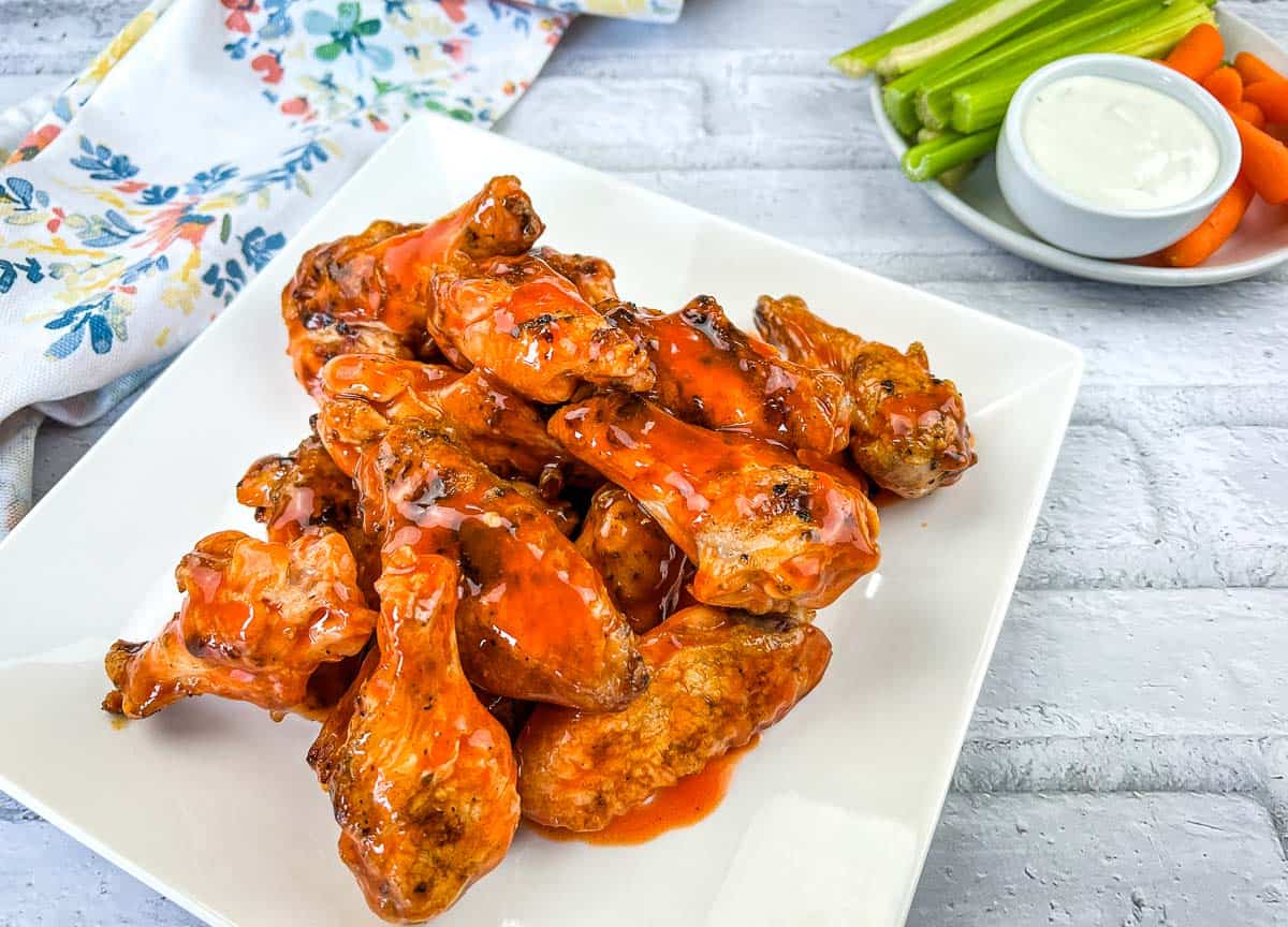 A square white plate with the best chicken wings, served alongside a small dish of ranch dressing, crisp celery sticks, and baby carrots.