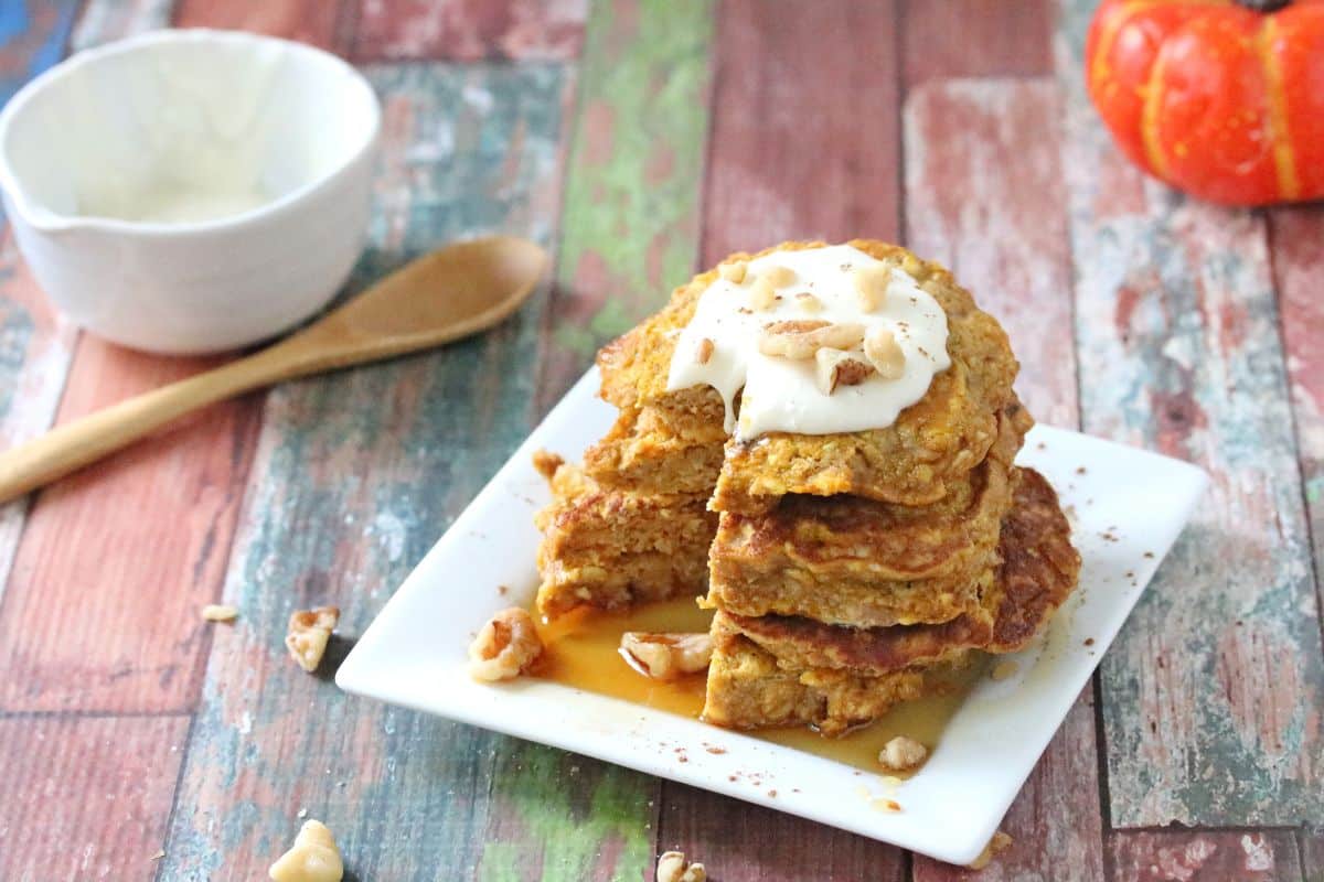 A stack of pancakes topped with cream and chopped nuts on a white plate, perfect for breakfast, with syrup and a wooden spoon beside a bowl on a rustic wooden table.
