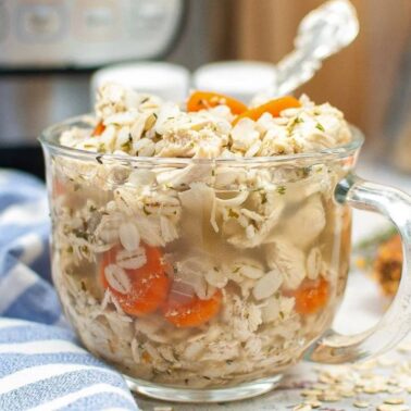 Clear glass mug filled with chicken soupโshredded chicken, oats, carrots, and herbsโserved with a spoon. A cozy takeout alternative and perfect for easy chicken recipes; kitchen items visible in the background.